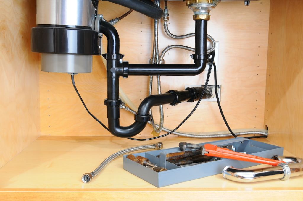Detail of the plumbing system under a modern kitchen sink, with a plumbers tool tray and equipment. Horizontal format.