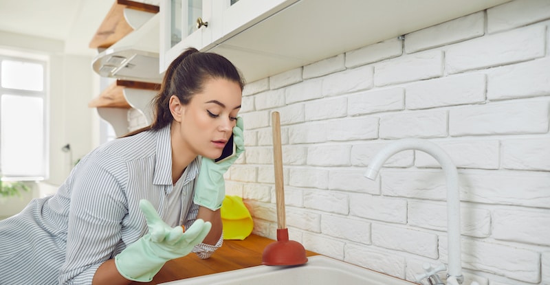 Young girl talking by the phone advising how to unclog a sink drain. Girl trying to clean sink using plunger standing in the kitchen at home. Housekeeping and household work concept.