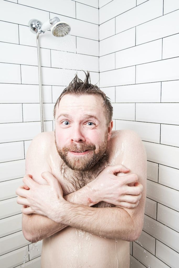 Caucasian bearded man with a funny expression on face feels the shock of taking a cold shower, he froze, tries to cover his body with hands and looks at the camera in the bathroom.