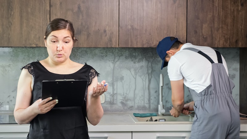 Dissatisfied housewife shocked by prices for plumbing services holding clipboard for signing. Male plumber fixes sink in modern kitchen