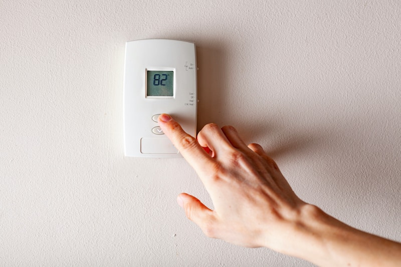 A woman is pressing the up button of a wall attached house thermostat with digital display showing the temperature. A concept image for electricity bill, heating, cooling, eco friendly, saving etc.