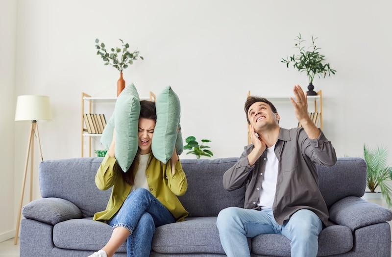 Couple sits on the sofa at home living room, covering ears due to loud noise from neighbors. They are annoyed by the renovation or loud music, showing frustration with the disturbance.