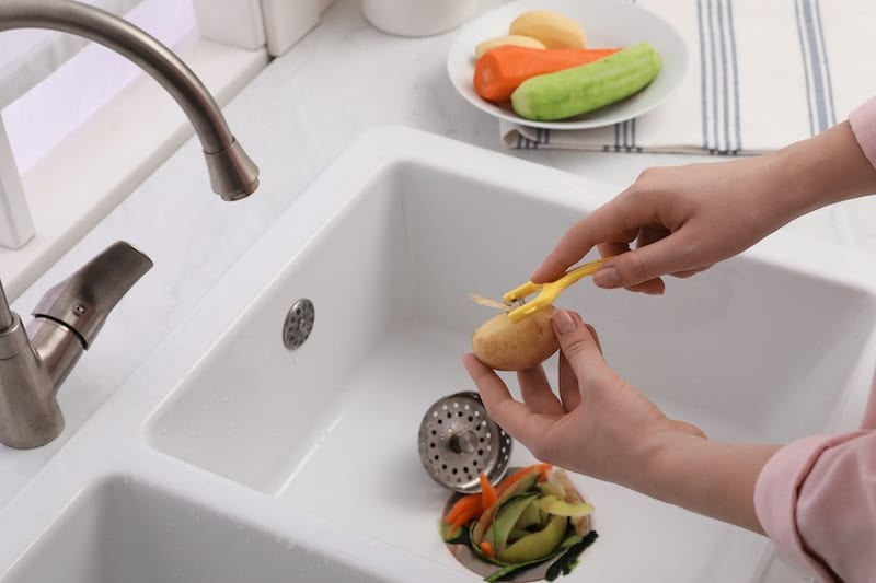 Woman peeling potato over kitchen sink with garbage disposal at home, closeup