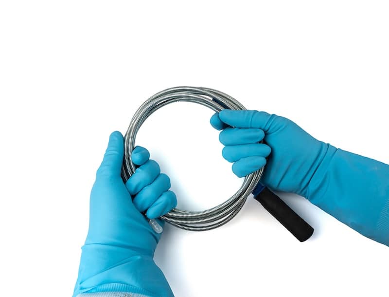 Metal drain auger tool isolated, plumbing snake or cable in hands in blue glove on white background, removing sink blockages instead of a chemical drain cleaner