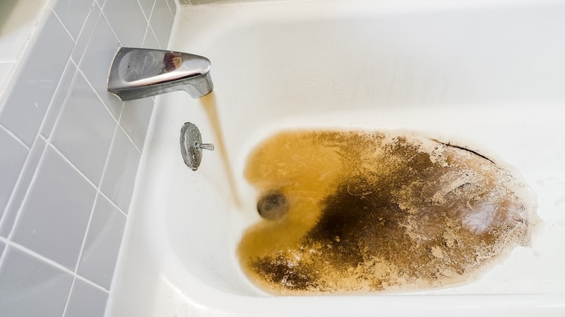 Brown water flows from the faucet into a bathtub, revealing dark sediment and discoloration in the water.