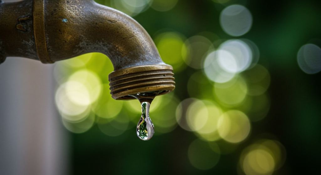 Close-up of an old brass faucet dripping water, set against a blurred green background. The focus is on the single water droplet.