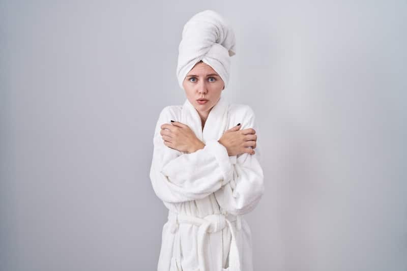 Blonde caucasian woman wearing bathrobe shaking and freezing for winter cold with sad and shock expression on face
