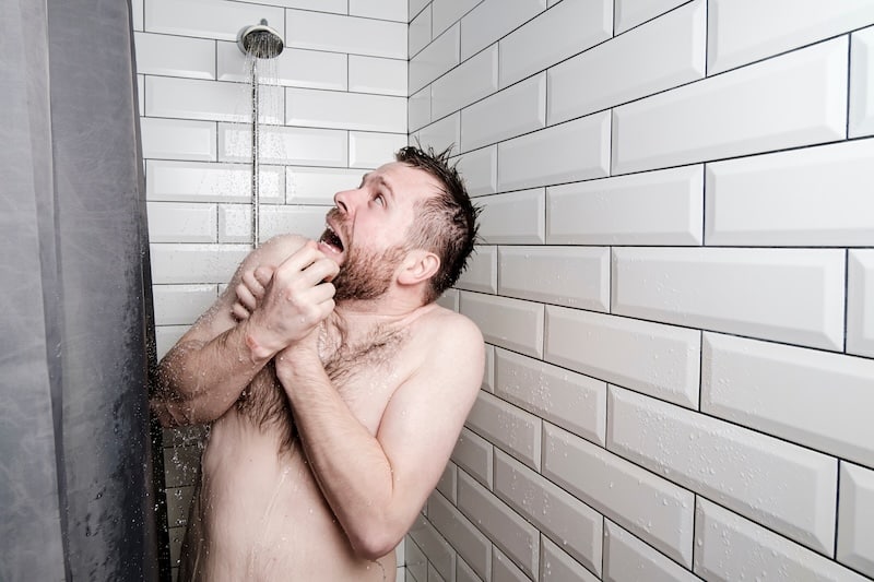 Shocked man looks at a watering can in the shower room, from which, unexpectedly, cold water is pouring, he freezes and trembles in the bathroom.