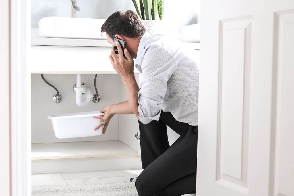 Sad Young Man Calling Plumber In Front Of Water Leaking From Sink Pipe