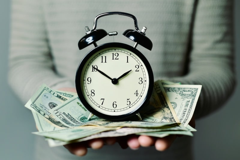 closeup of a young caucasian man with an alarm clock and many US dollar banknotes in his hands, depicting the idea that time is money