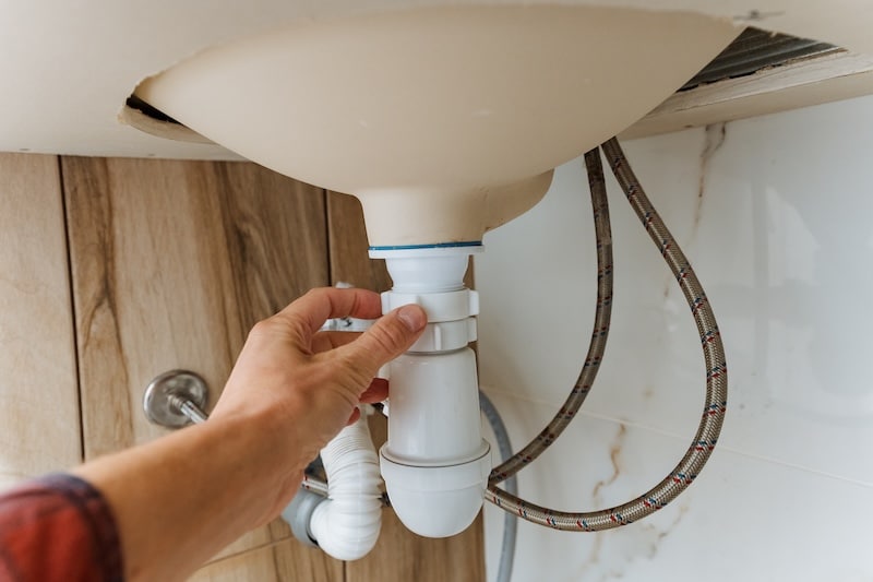 hand reaching under wooden sink basin to tighten trap and check connections, visible braided hoses and cabinet base, handson plumbing maintenance scene for home repair and service content